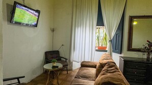Ein Wohnzimmer mit braunem Sofa, einem kleinen Tisch, einem an der Wand befestigten Fernseher und einem Fenster mit Vorhängen.