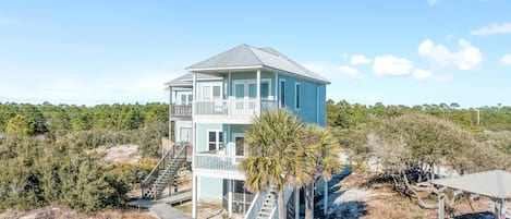 A two-story beachfront house with a balcony, a wooden deck, and a small patio area.