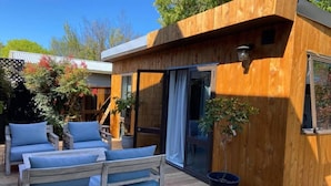 Una cabaña de madera con puerta corredera de vidrio, un pequeño patio con banco y cojines de madera, y plantas en macetas.