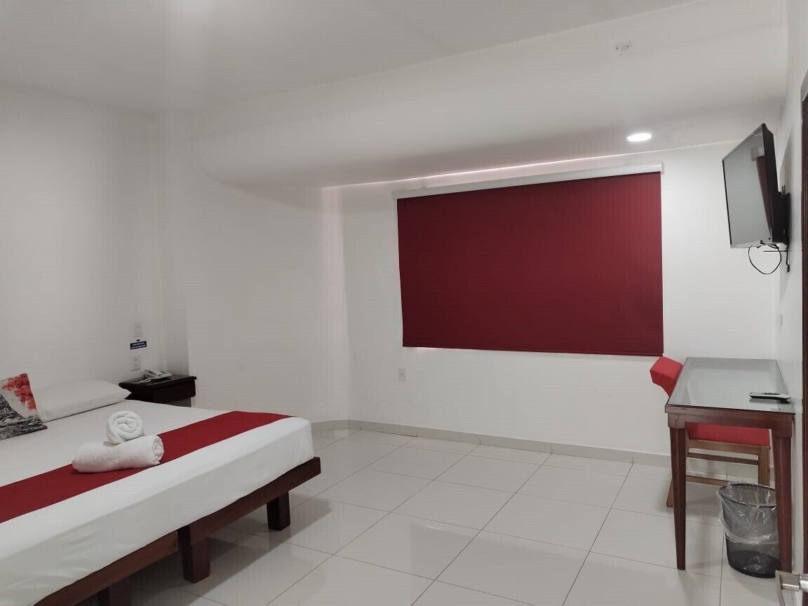 A hotel room with a bed, a desk, a chair, a TV, and a window with a red blind.