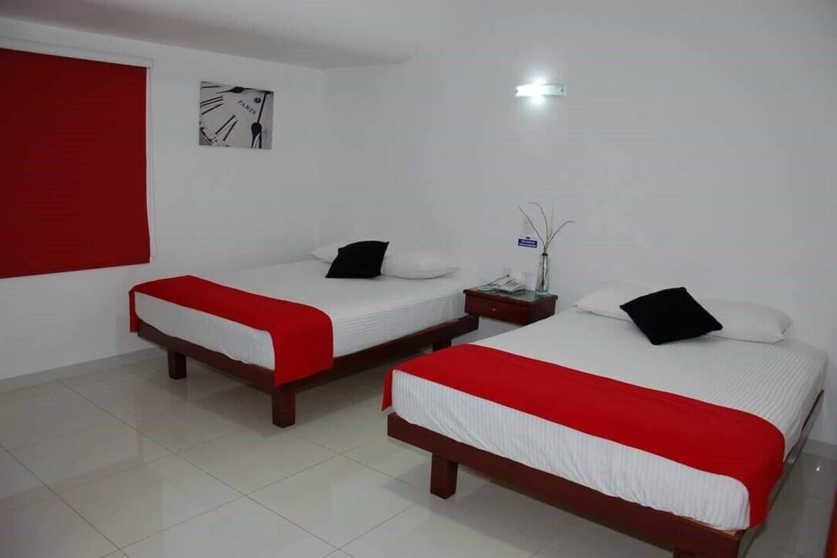 A hotel room with two beds, each with a red bedspread and white linens, a nightstand with a lamp, a wall clock, and a small potted plant.