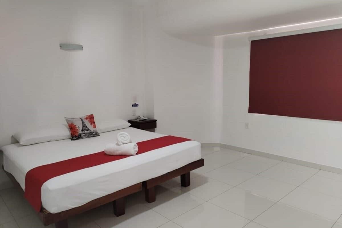 A neatly made bed with white and red bedding, a small bedside table with a lamp, and a plain white wall with a red accent.