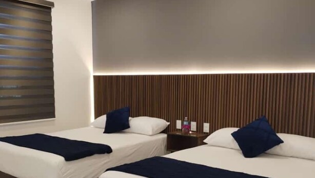 Une chambre d’hôtel avec deux lits, chacun recouvert d’une courtepointe et doté d’oreillers bleu foncé, ainsi qu’une tête de lit en bois et une fenêtre avec des stores.