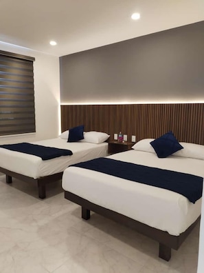 A hotel room with two beds, each with a dark blue bedspread and pillows, a wooden headboard, and a window with blinds.