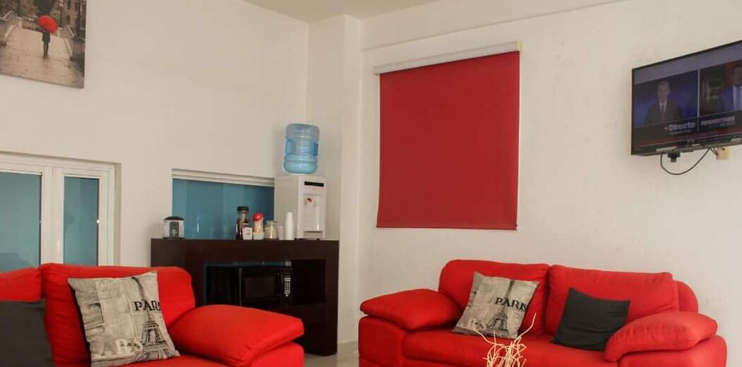 Uma sala de estar com dois sofás vermelhos, uma televisão na parede e um dispensador de água em um armário.