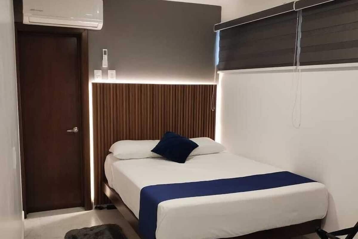 A modern hotel room with a bed, a bedside table, a small stool, a wall-mounted air conditioner, and a window with blinds.