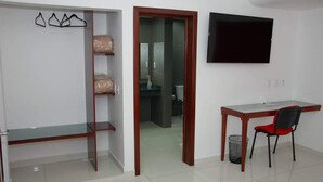 Une pièce comprenant un bureau, une chaise, un téléviseur, une armoire et une salle de bain visible à travers une porte ouverte.