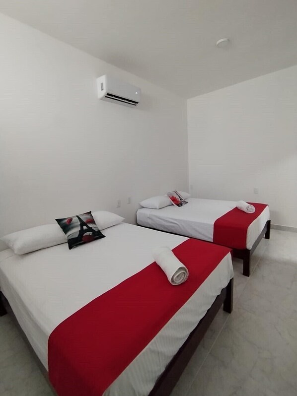 A hotel room with two beds, each with white bedding and red accents, a white pillow with a red design, and a white air conditioning unit on the wall.