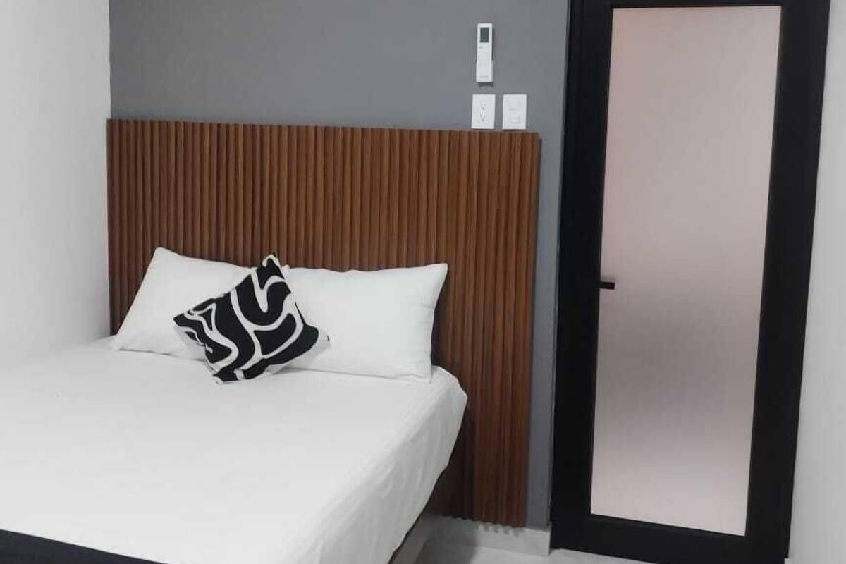 A modern hotel room with a bed, a wooden headboard, a white air conditioning unit, a door, and a small rug.