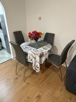 A dining area with a table covered by a floral tablecloth, surrounded by four chairs with metal legs. A vase with flowers is placed on the table. - Charming Escape: 2BR Home with Parking (Aveley)