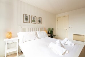 Un dormitorio con una cama blanca, una mesita de noche con lámpara e impresiones botánicas enmarcadas en la pared.