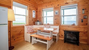 A cozy cabin interior with wooden walls, a white bench, a table, a lamp, and a fireplace. - Windy Dreams: an idyllic, cozy seaside PEI cottage (Kensington)