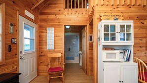 A wooden cabin interior with a cozy fireplace, a wooden chair with a red cushion, a microwave, and a cabinet with dishes. - Windy Dreams: an idyllic, cozy seaside PEI cottage (Kensington)