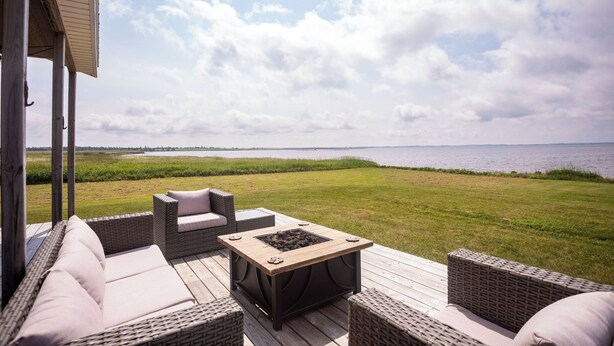 An outdoor deck with wicker furniture, a square table with a fire pit, and a view of a body of water and grassy fields. - Windy Dreams: an idyllic, cozy seaside PEI cottage (Kensington)