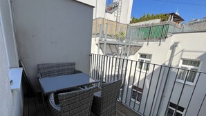 Een balkon met rieten meubels, een metalen hekwerk en uitzicht op omliggende gebouwen.