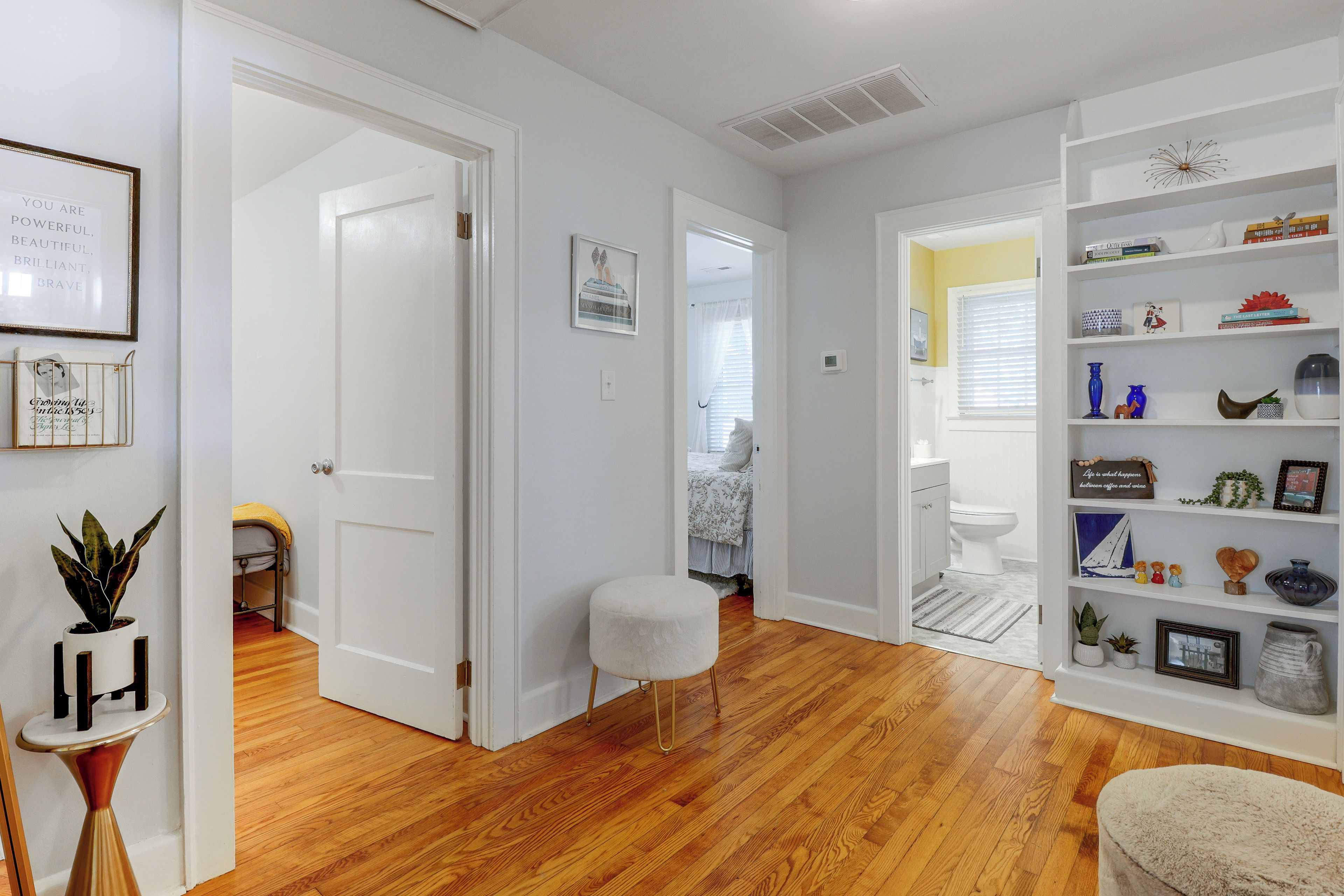 Un couloir avec une porte blanche donnant sur une chambre, un petit tabouret blanc et une bibliothèque contenant divers objets.