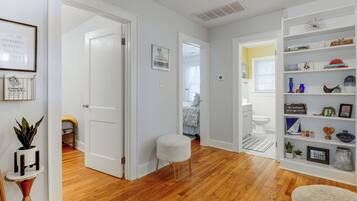 Un couloir avec une porte blanche donnant sur une chambre, un petit tabouret blanc et une bibliothèque contenant divers objets.
