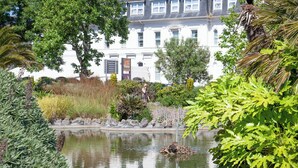 Un edificio con facciata bianca e numerose finestre, circondato da vegetazione rigogliosa e uno stagno.