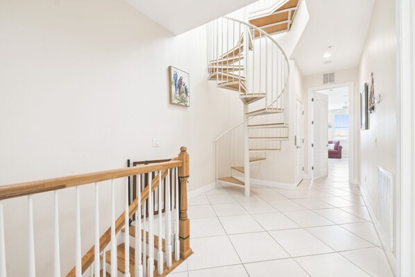 A modern staircase with a wooden handrail and white railings, leading to a room with white tiled flooring and a door at the end.