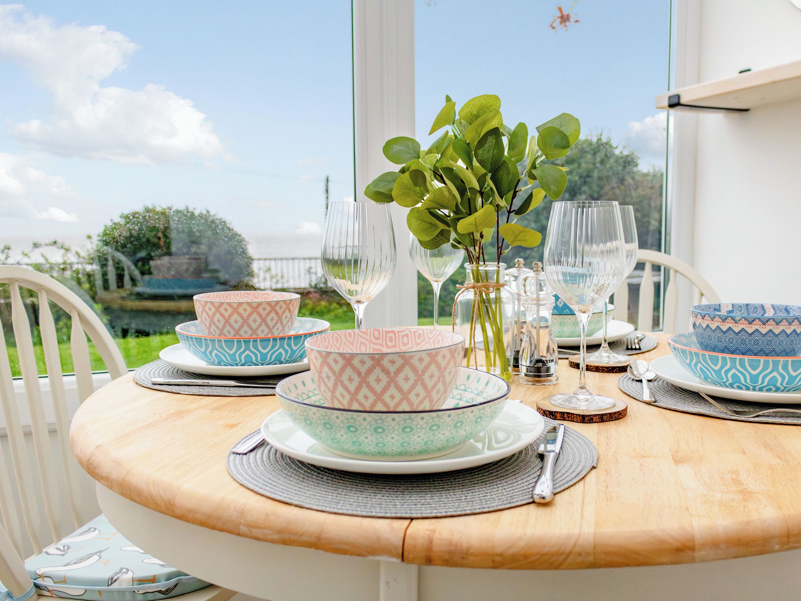 A round dining table with a view of the outdoors, set with patterned bowls, wine glasses, and a vase with green leaves.