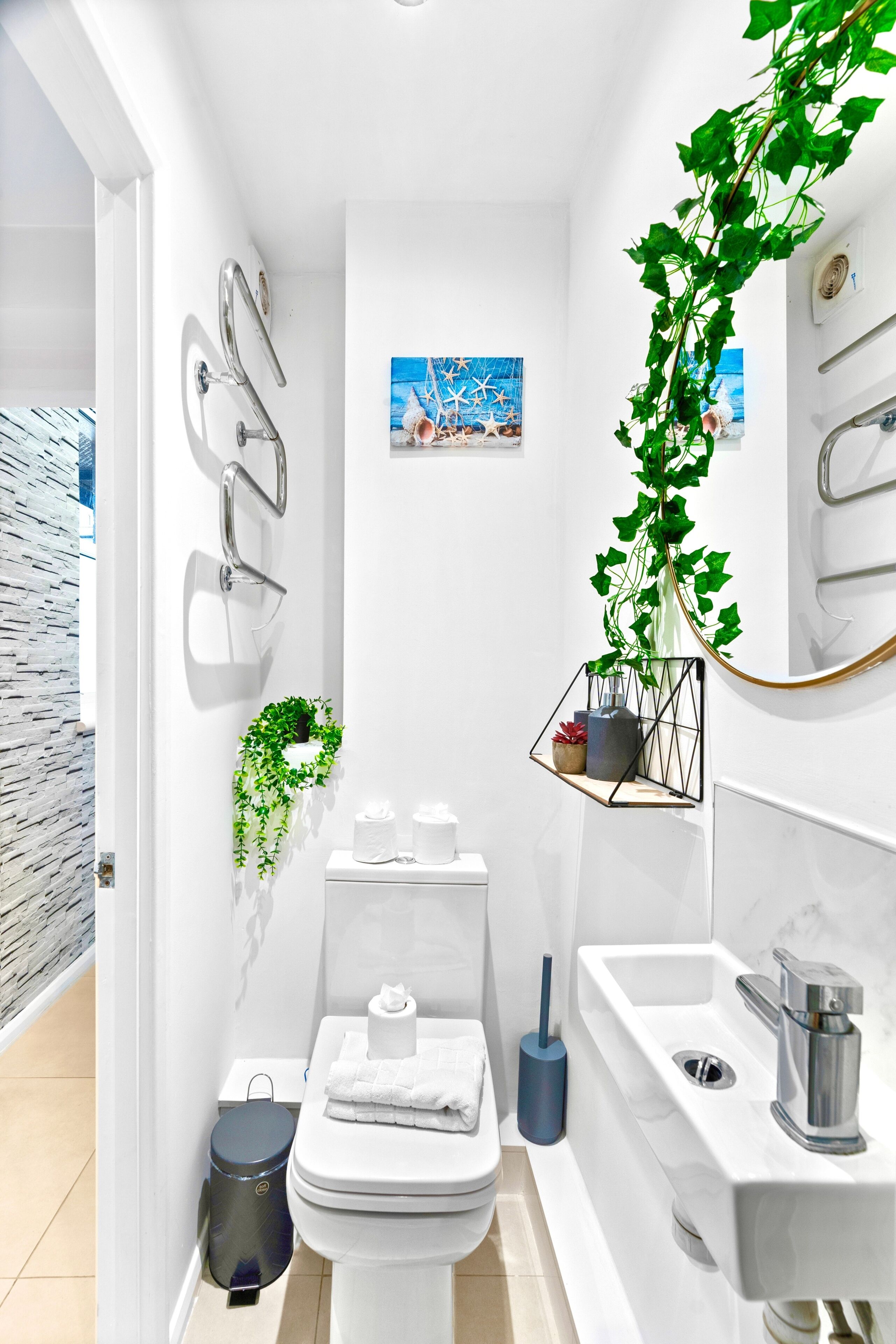 Un baño moderno con inodoro, lavabo y zona de ducha. Hay plantas y un cuadro en la pared.