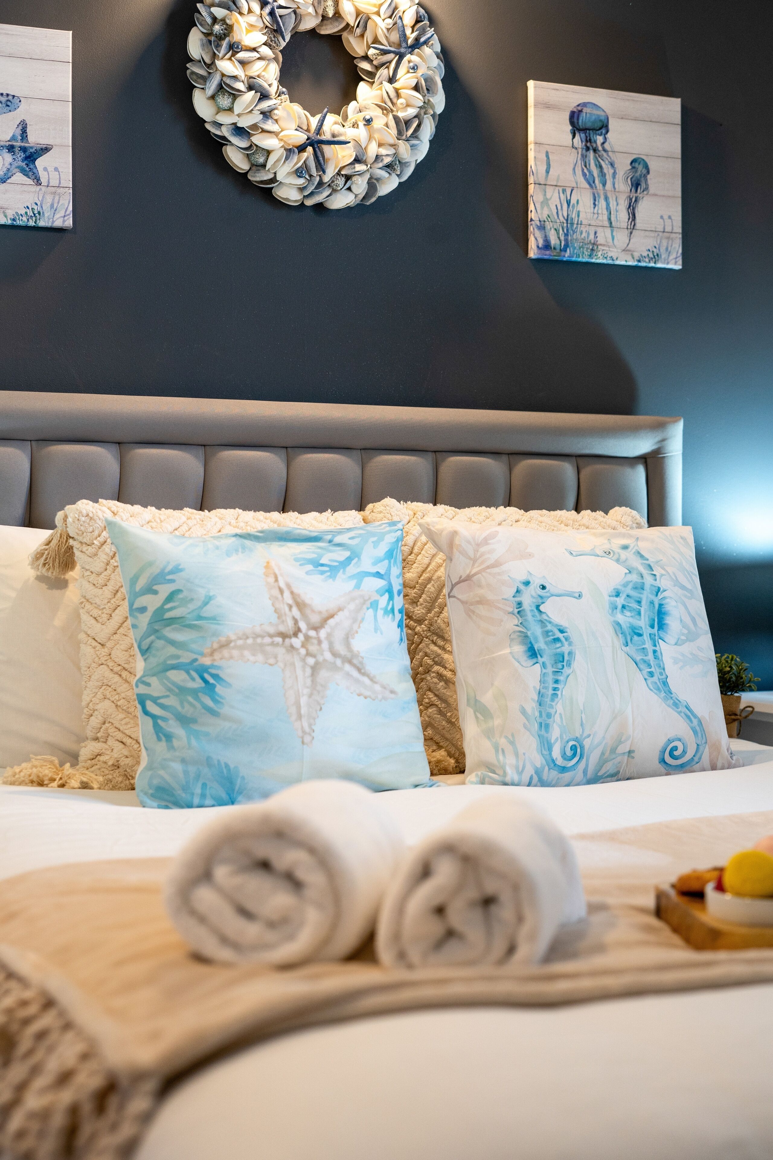 A neatly made bed with a starfish and seahorse pillow design, a towel, and a fruit bowl on the side.