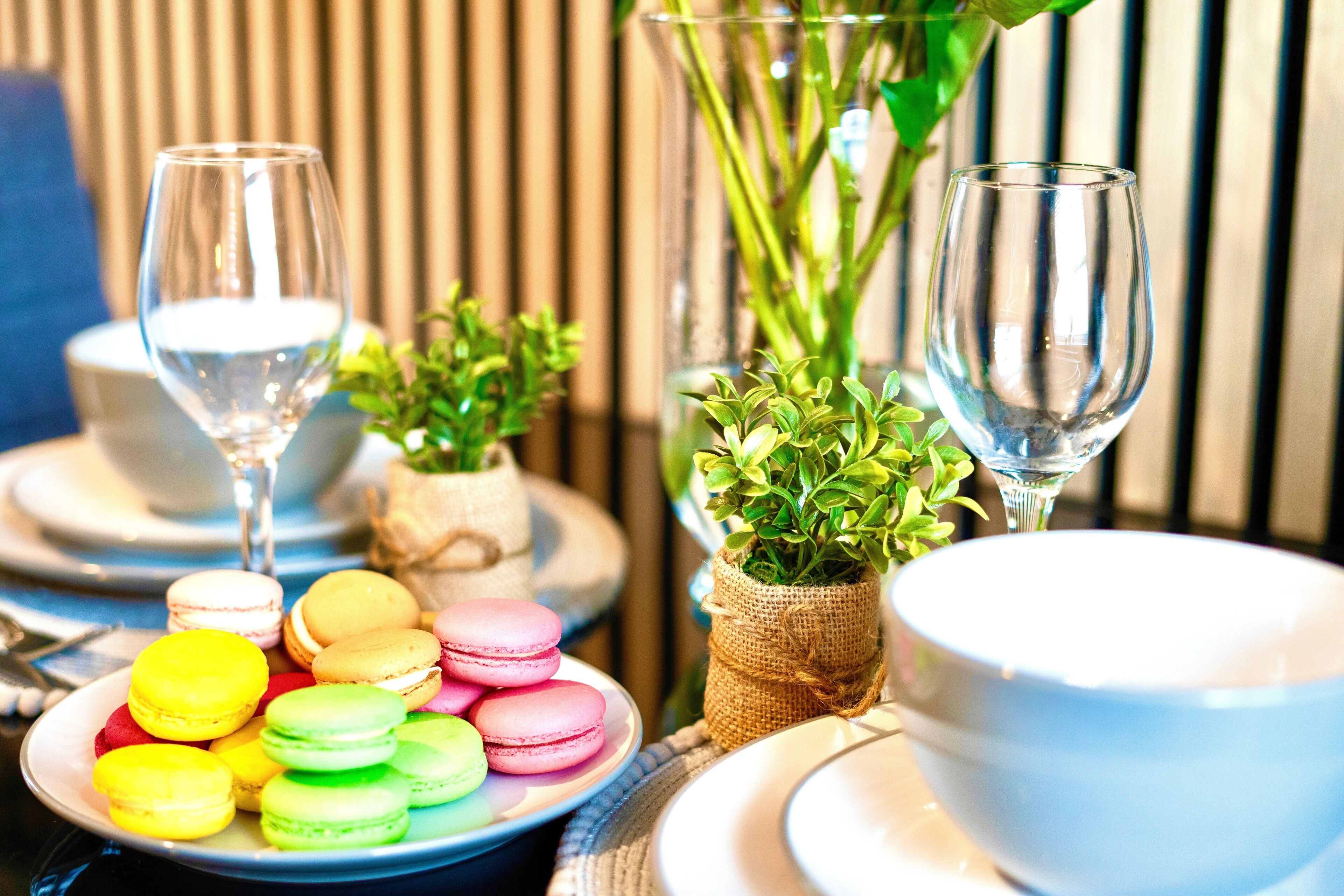 Une table dressée avec des macarons aux couleurs vives, deux verres à vin et de petites plantes en pot.