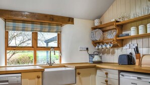 A kitchen with wooden cabinets, a built-in dishwasher, a window with a plaid curtain, and a shelf with dishes and a kettle. - Lower Axford Cottage (Callington)