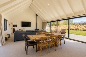 A spacious living area with a large sofa, a dining table with chairs, a flat-screen TV mounted on the wall, and a view of the outdoors through large windows. - The Big House, Twizel (Twizel)