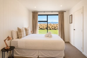 A bedroom with a large bed, a bedside lamp, a view of a grassy field through a window, and a door to the right. - The Big House, Twizel (Twizel)