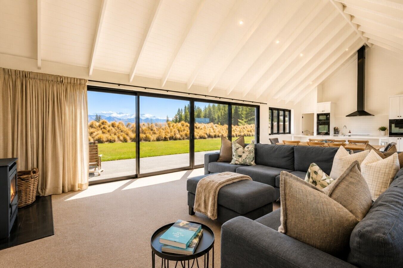 A modern living room with a sofa, a coffee table, and a fireplace. There is a large window offering a view of a grassy field and mountains.