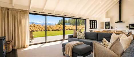 A modern living room with a sofa, a coffee table, and a fireplace. There is a large window offering a view of a grassy field and mountains.