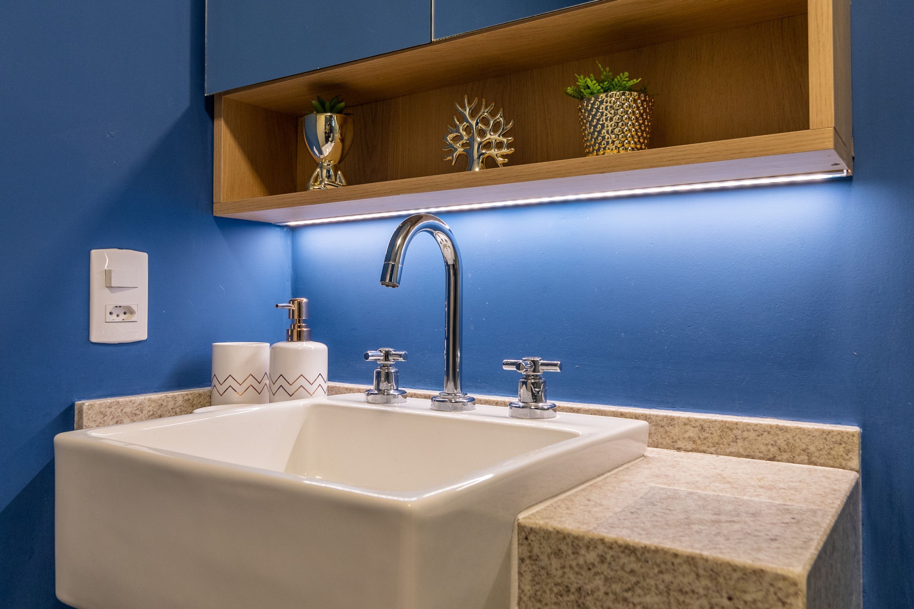 A bathroom with a white sink, two chrome faucets, a wooden shelf above the sink, and a small plant.