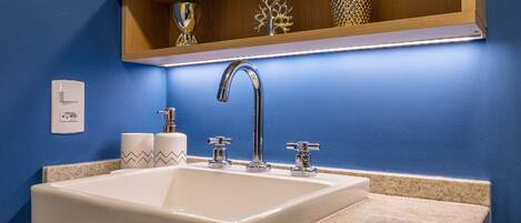 A bathroom with a white sink, two chrome faucets, a wooden shelf above the sink, and a small plant.