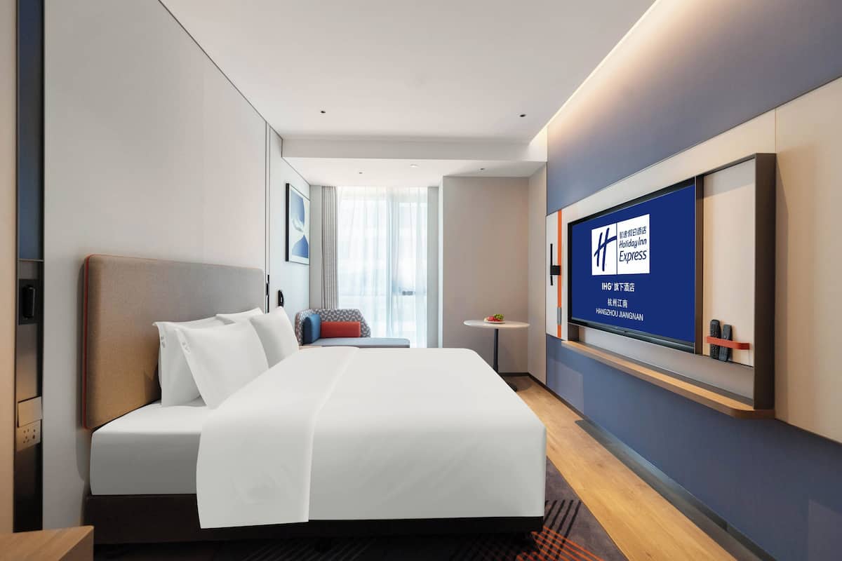 Ein modernes Hotelzimmer mit einem großen Bett, einem Nachttisch, einem kleinen runden Tisch und einem Fernseher, der das Holiday Inn Express Logo zeigt.
