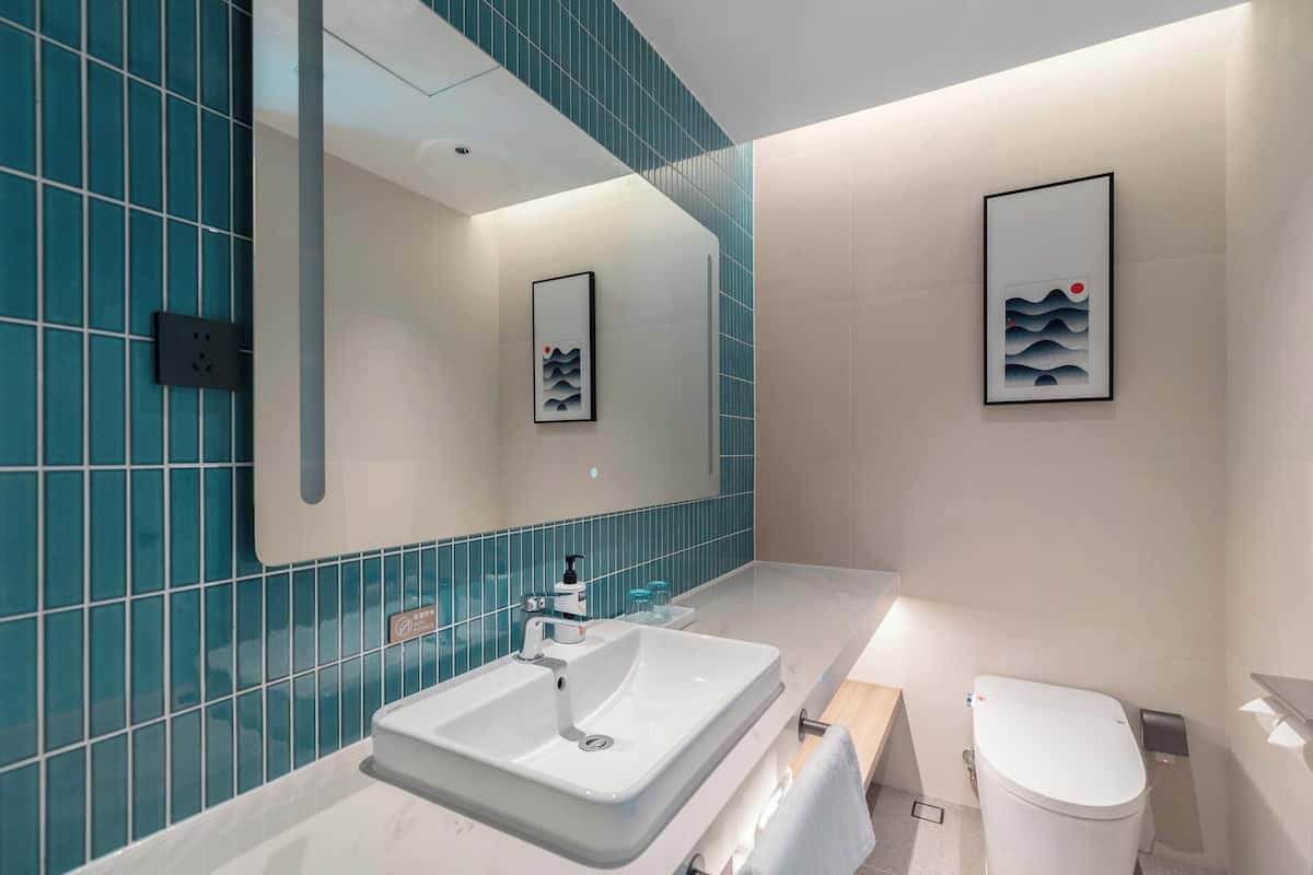Ein modernes Badezimmer mit einem weißen Waschbecken, einer Toilette und Kacheln in Türkis. An den Wänden befinden sich Bilder in Rahmen und ein Spiegel über dem Waschbecken.