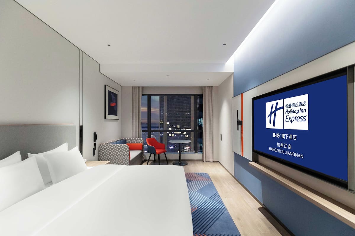 Ein modernes Hotelzimmer mit einem großen Bett, einem kleinen Tisch, einem roten Sessel und einem großen Fenster mit Blick auf die Stadt.