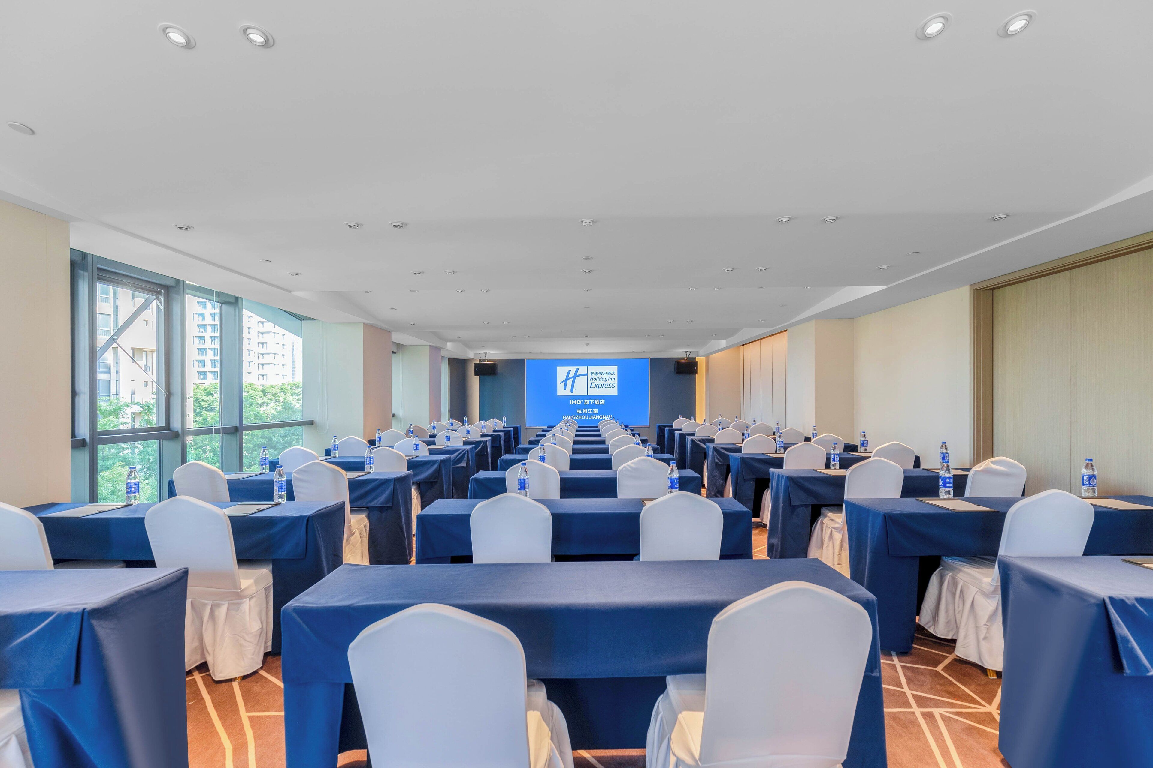 A conference room with rows of tables and chairs, large windows, and a presentation screen.