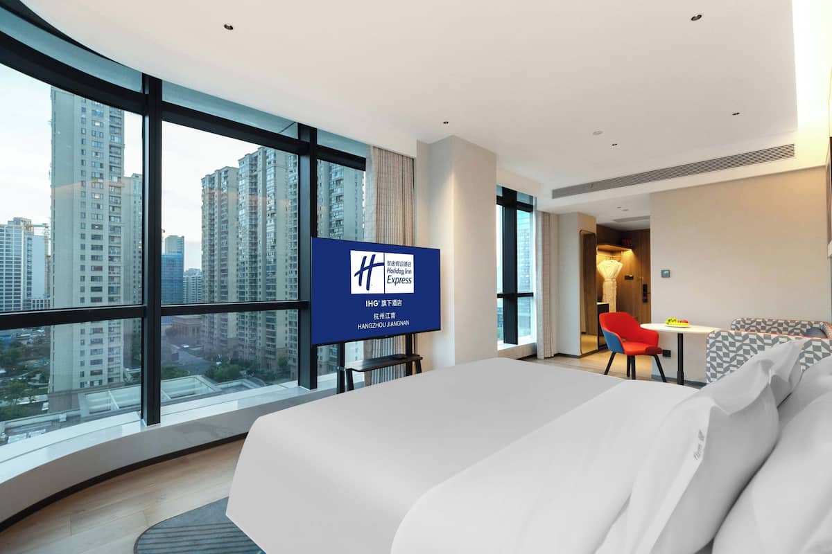 Ein modernes Hotelzimmer mit einem großen Bett, einem Schreibtisch und einem Sessel. Ein großes Fenster bietet Blick auf die Stadt. Ein Schild an der Wand zeigt den Namen und den Standort des Hotels.