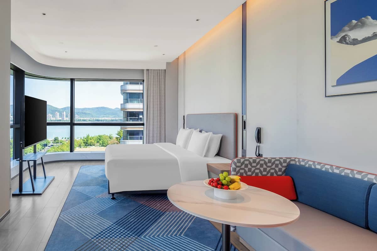 Ein modernes Hotelzimmer mit einem großen Bett, einer Couch, einem Esstisch mit Obst, einem Fernseher und Blick auf die Stadt.