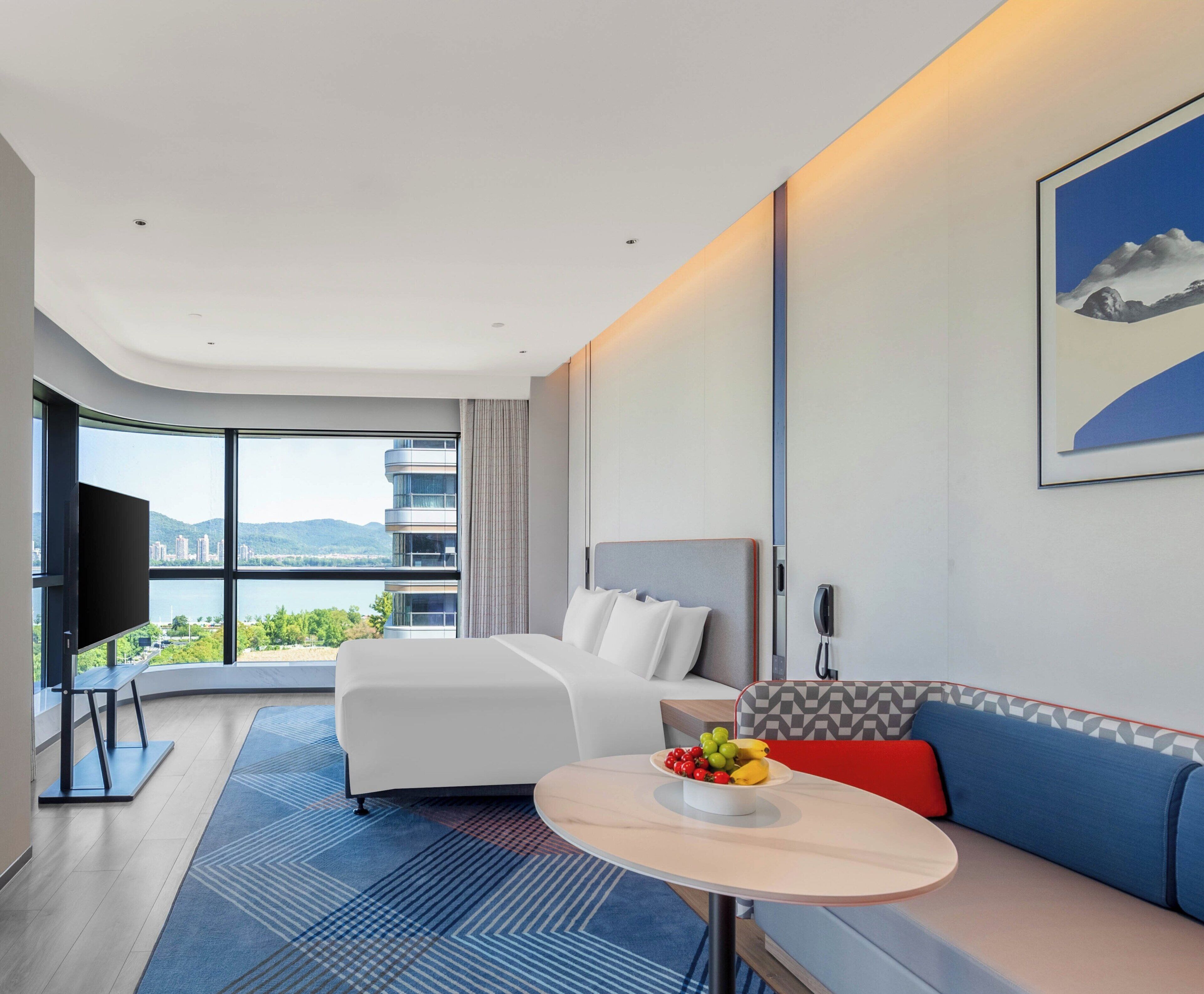 Habitación de hotel moderna con una cama grande, un sofá, una mesa de comedor con fruta, un televisor y vistas a la ciudad.