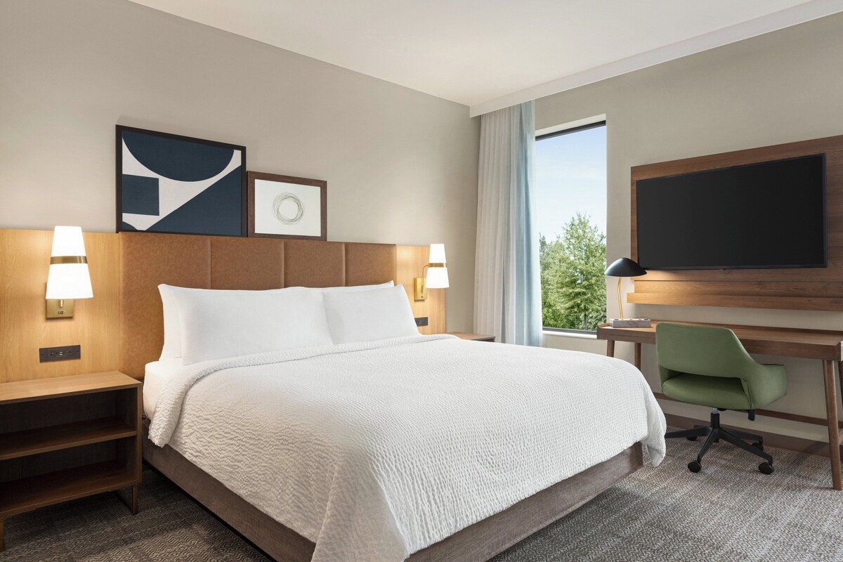 A hotel room with a large bed, a desk with a chair, a flat-screen TV, and artwork on the wall.