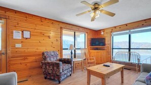 Interior de una cabaña de madera con ventilador de techo, televisor, sillón estampado, mesa de madera y balcón con vista a las montañas.