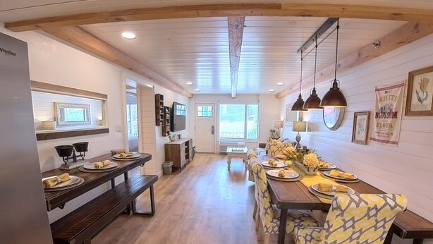 A spacious dining area with wooden tables and benches, a kitchen area with a refrigerator and a television, and a large window providing natural light. - "Farmhouse Chic: Poolside Luxury" Sleeps 16 (Pigeon Forge)