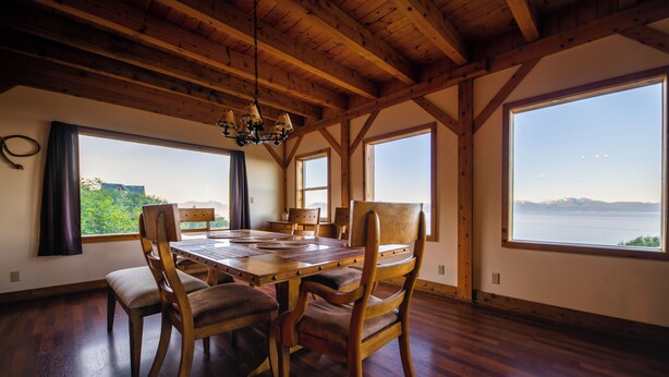 A dining area with a wooden table and chairs, large windows with a view of the mountains, and a wooden ceiling. - Cozy lodge near town w Stunning Views & Fireplace (Homer)