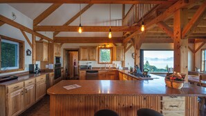 A spacious kitchen with wooden cabinets, a large island with stools, and a view of the mountains through the windows. - Cozy lodge near town w Stunning Views & Fireplace (Homer)