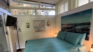 Un dormitorio con una cama, televisión, un cuadro y una ventana con vista a árboles.