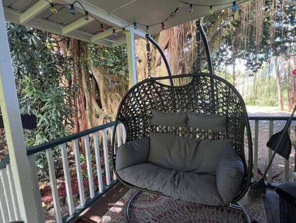 A hanging chair with cushions in a covered outdoor area with a view of trees and a pathway.