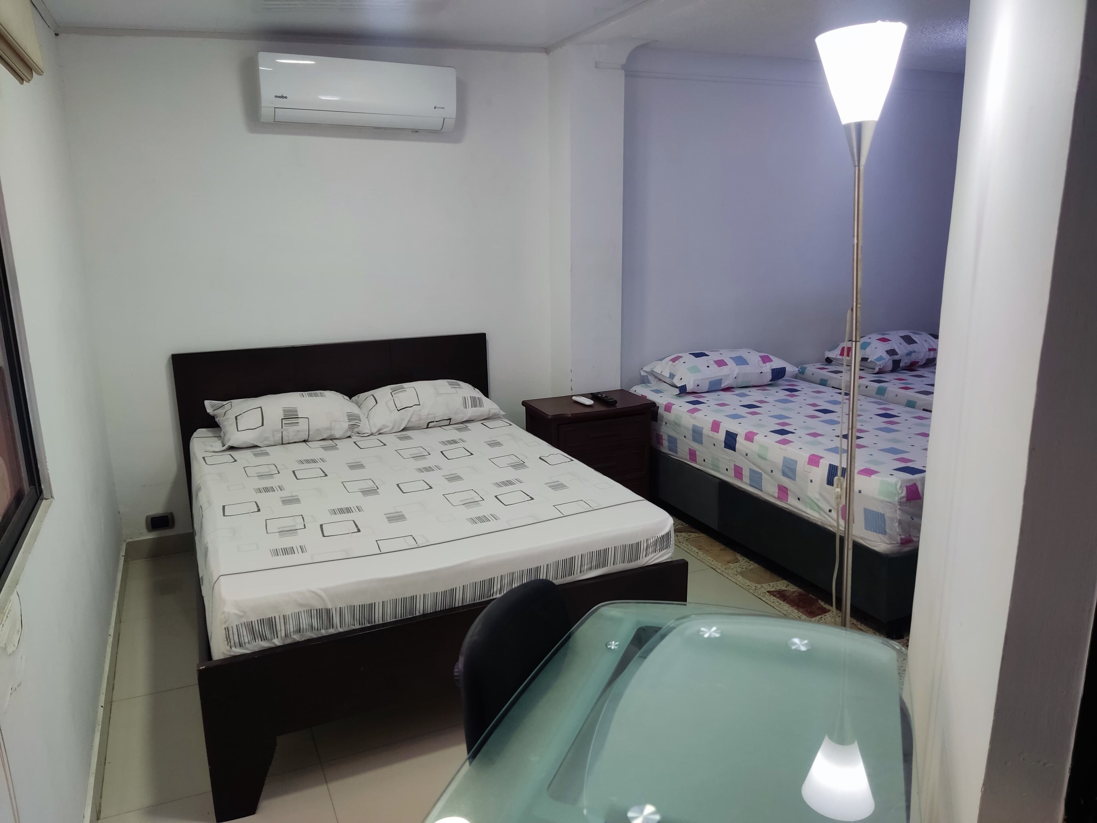 A bedroom with a bed, bedside tables, a desk, a lamp, and an air conditioner.