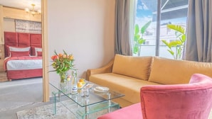 Une chambre d’hôtel moderne avec un lit rouge, un canapé beige, une table basse en verre et une vue sur la ville grâce à de grandes fenêtres.